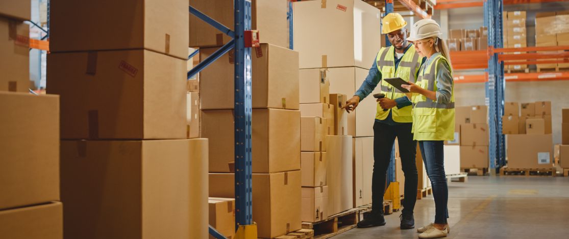 Wo workers checking boxes in a large warehouse wearing safety gear